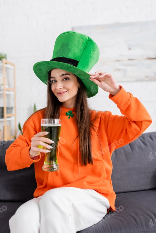 Preview: Positive young woman in green hat holding beer while sitting on couch