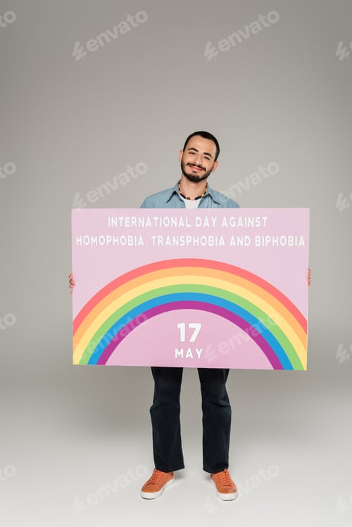 Visualização: homem homossexual sorrindo e segurando cartaz com o Dia Internacional contra a Homofobia, Transfobia e