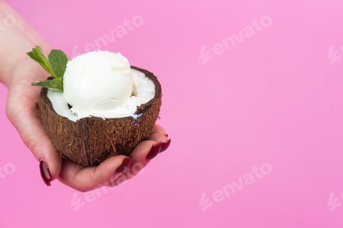Preview: Vanilla ice cream balls in a fresh coconut half, decorated with mint leaves in your hand on a pink