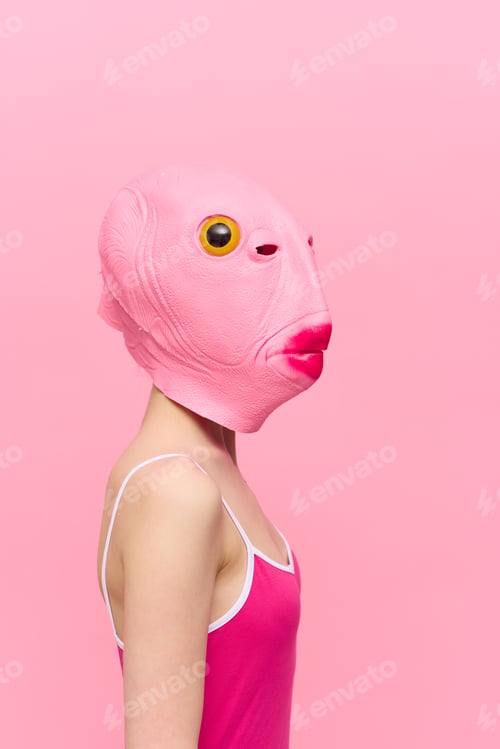 Preview: A woman in a pink fish head mask stands in profile against a pink background and looks into the