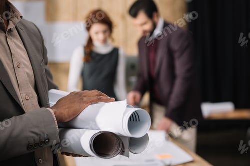 Preview: cropped shot of architect holding rolled plans with blurred colleagues working on background