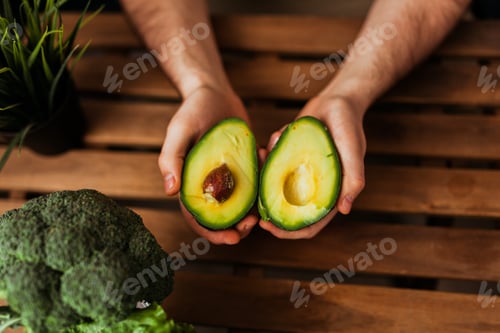 Preview: young man holding cut avocado over wooden table