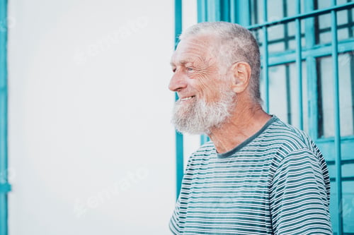 Preview: Smiling elderly man with white beard wearing striped shirt standing by blue window frame