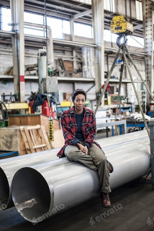 Preview: Black woman sitting next to a lift with aluminum pipes in a sheet metal factory.