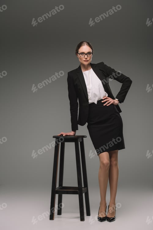 Preview: beautiful businesswoman in eyeglasses posing near chair on grey background