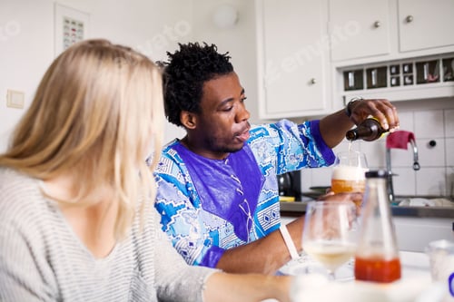 Preview: Young woman looking at friend pouring beer in glass at home