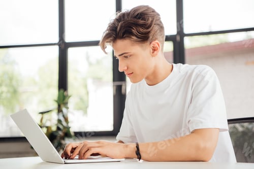 Preview: focused teen boy in white t-shirt using laptop at home
