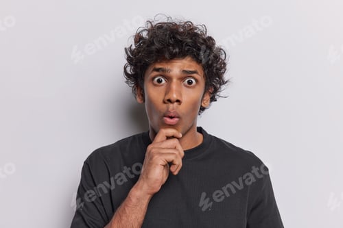 Preview: Photo of astonished curly haired Hindu man keeps hand on chin stares impressed reacts to