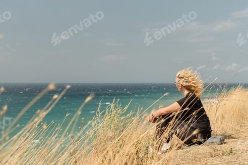 Preview: An adult woman sits on a high bank overlooking the ocean, her hair blowing in the wind