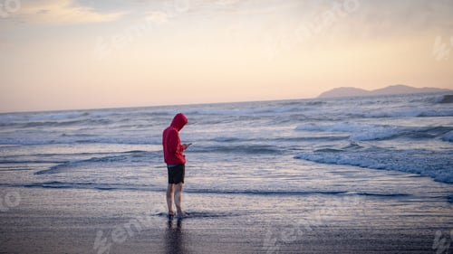 Preview: Young man in a red hoodie standing on a beach at sunset while looking at his phone