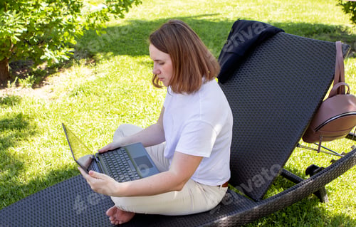 Preview: young girl sits on a sun lounger in a city park and works on a computer