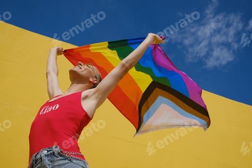 Preview: Cheerful non-binary person holding up and waving an lgbt pride flag outdoors.