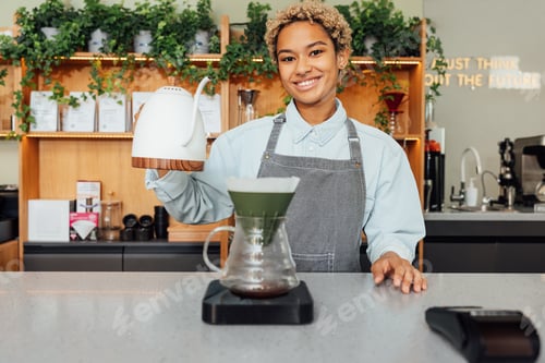 Preview: Smiling female barista holding a kettle while standing at a counter in coffee shop