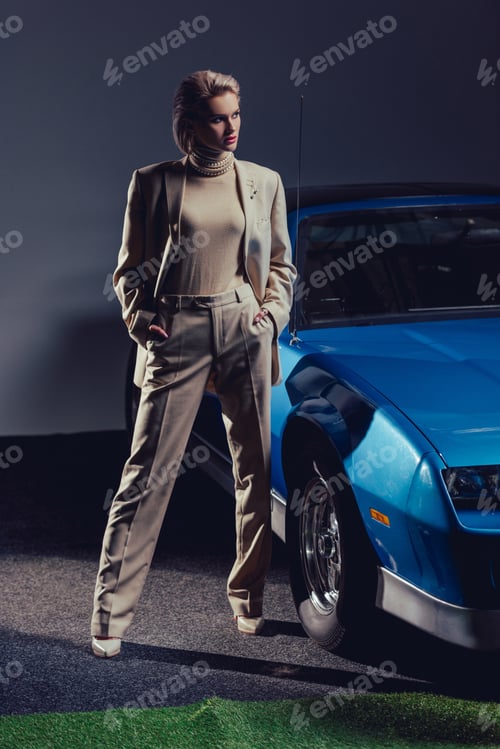 Preview: attractive and stylish woman in suit with hands in pockets standing near retro car