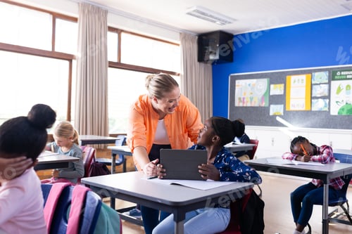 Preview: Female teacher helping african american girl with tablet in classroom, other students in school