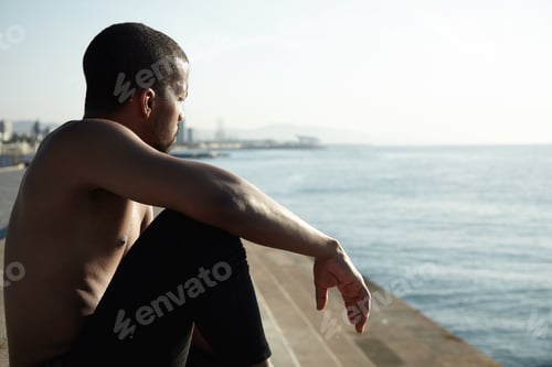 Preview: Handosme dark-skinned sportsman with muscular athlete body sitting on sidewalks at ocean, watching,