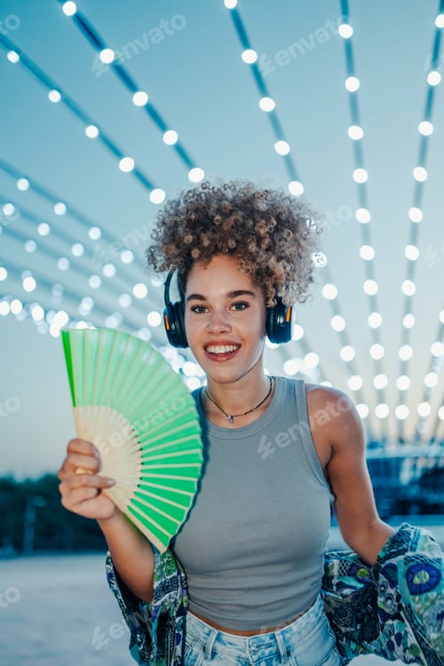 Preview: Young woman listening music with headphones and cooling with hand fan at summer festival
