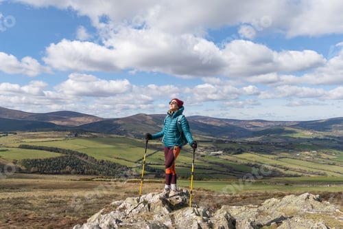 Preview: Adult female hiker with trekking poles standing on a rock under a blue cloudy sky