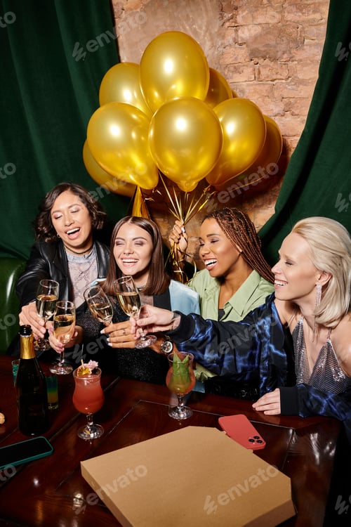 Preview: multiethnic girlfriends clinking champagne glasses near golden balloons in bar, birthday party