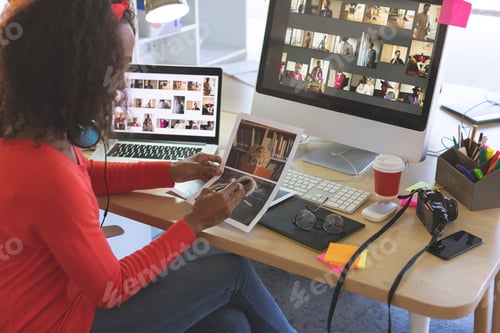 Preview: Side view of young mixed-race female graphic designer looking photographs at desk in a modern office