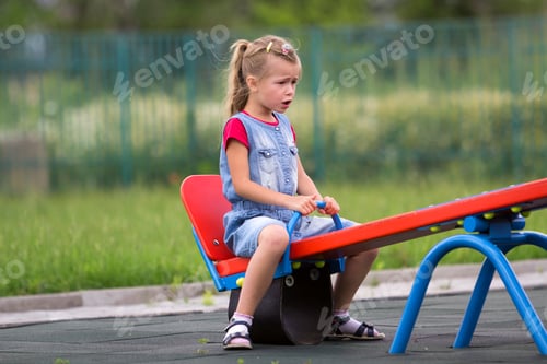 Preview: Small cute young blond child girl sits moody, angry and unsatisfied on see- saw swing on warm sunny