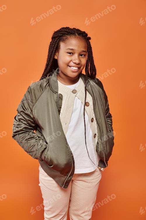 Preview: merry preadolescent african american girl with braids posing with hands in pockets smiling at camera