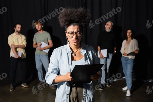 Preview: frowning african american woman holding clipboard and looking at camera near multicultural actors