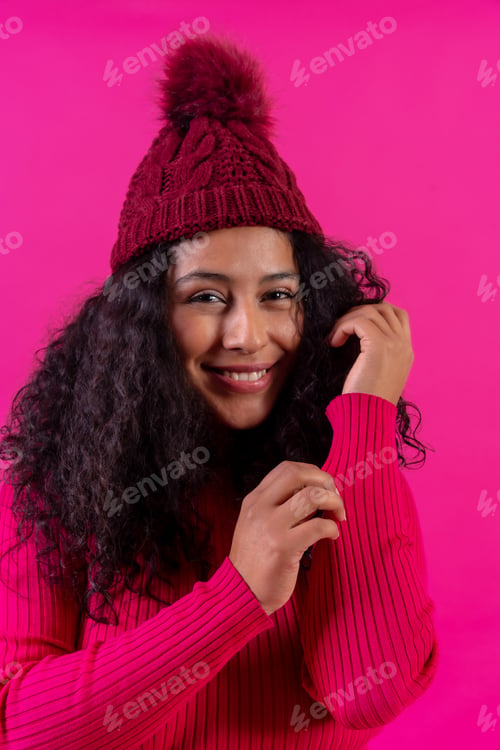 Preview: Curly-haired woman in a wool cap on a pink background portrait smiling, studio shot