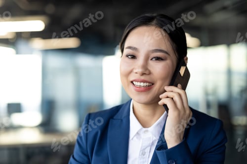 Preview: Asian businesswoman in blue suit smiling while talking on smartphone in modern office environment