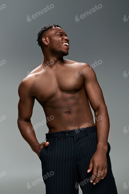 Preview: cheerful african american man with shirtless muscular body and hand in pocket looking away on grey