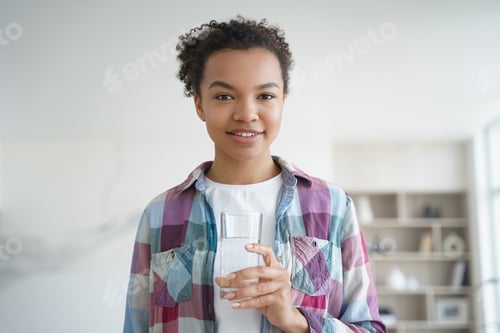 Preview: African american girl holds glass of purified water standing at home. Healthy lifestyle, wellness