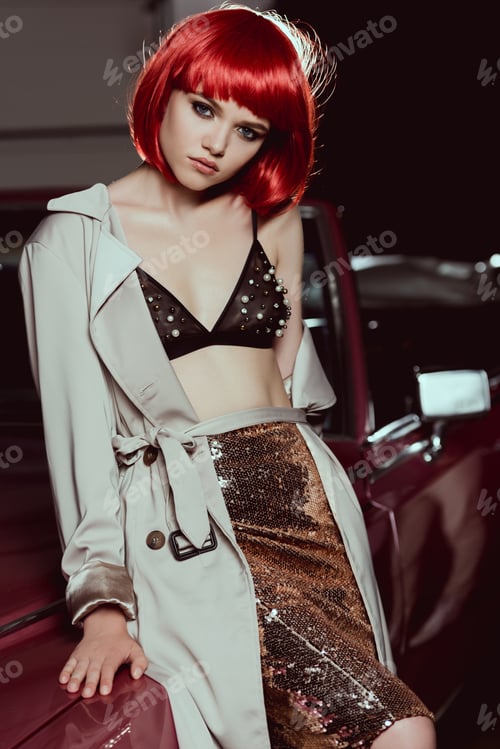 Preview: beautiful young woman in red wig, bra and trench coat sitting on classic car and looking at camera