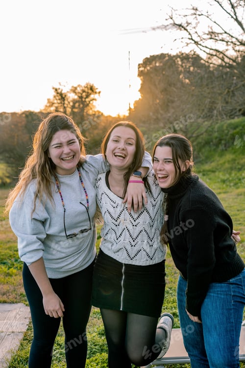 Preview: Portrait of three young women laughing loudly in a park at sunset. Friendship concept