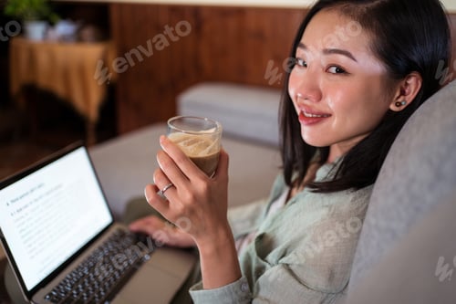 Preview: Asian freelancer typing on laptop on couch while drinking latte