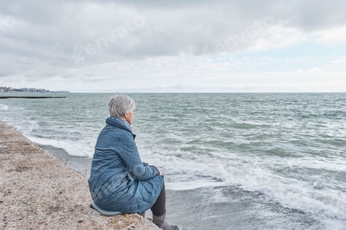 Preview: Senior woman sitting on sea coast on cold cloudy day.