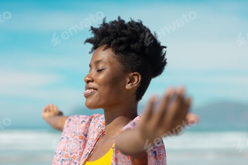 Preview: Smiling african american young woman with eyes closed and arms outstretched against sky on sunny day