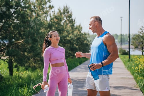 Preview: Man and woman in colorful athletic wear, laughing together, holding water bottles, on a sunny day in
