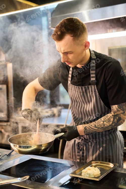 Preview: Young Caucasian Man Cooking Food in Professional Kitchen While Stirring Pan