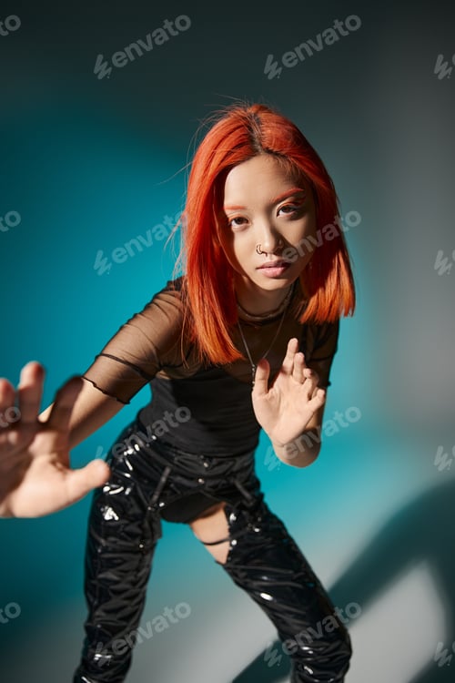 Preview: asian woman with piercing and red dyed hair gesturing and looking at camera on blue background