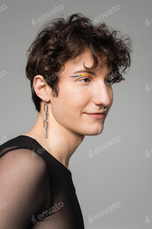 Preview: portrait of smiling bigender model with rainbow eye liner and silver accessories isolated on grey
