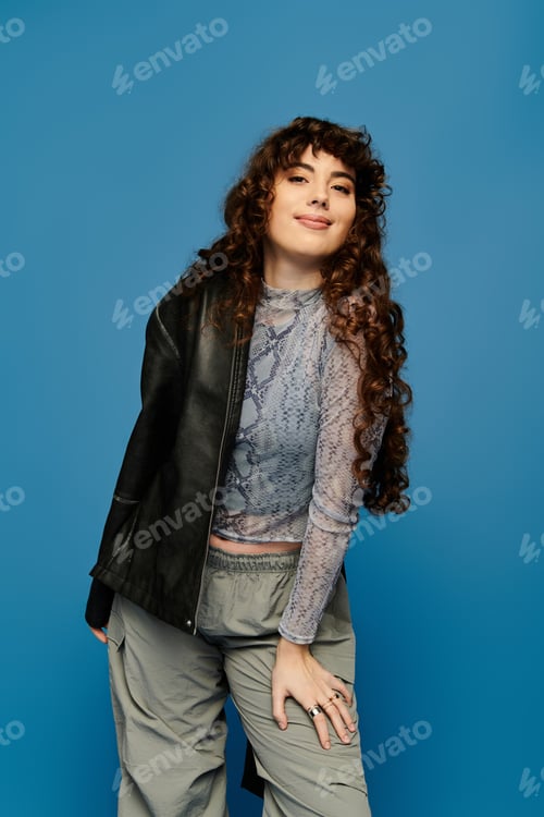 Preview: Stylish young woman expressing emotion against a vibrant blue backdrop with a trendy outfit
