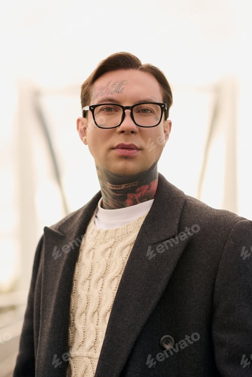 Preview: Stylish young man with tattoos showcases modern fashion against a blurred background
