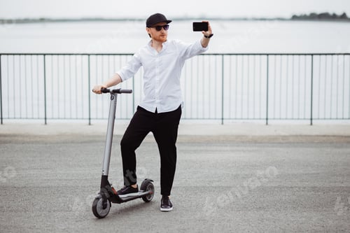 Preview: Modern man in stylish outfit making selfie while standing at the street with electric scooter