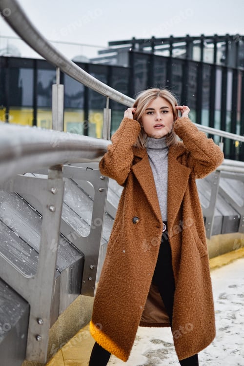 Preview: Young woman straightens her hair outdoors in winter while standing on the bridge of a big city.
