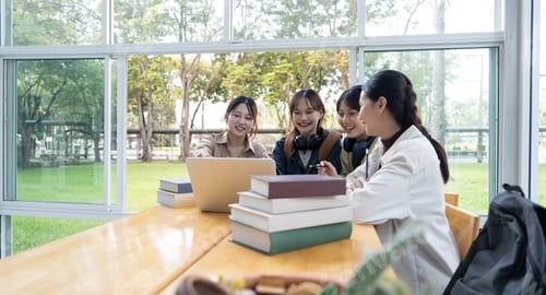 Preview: University students sitting together at table with book and laptop. Happy young people doing group