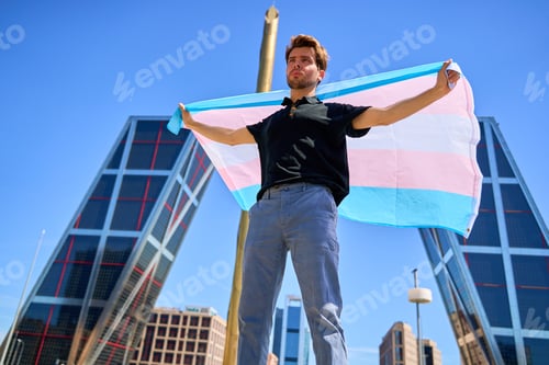 Preview: A man holding a transgender flag in front of a building. The flag is large and colorful.