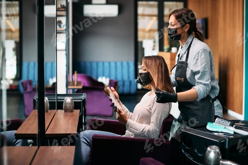 Preview: Young woman and her hairdresser wearing face masks while choosing hair color at the salon.