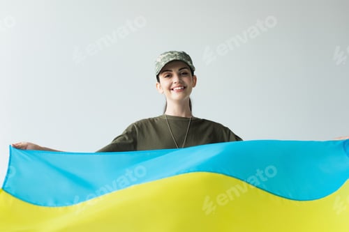 Preview: Positive soldier in cap holding ukrainian flag isolated on grey