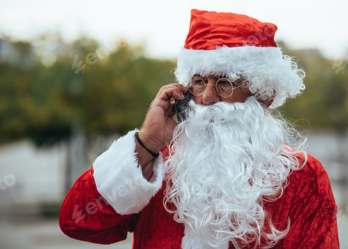 Preview: Stock photo of Santa claus talking to a mobile phone with a park behind unfocused. Christmas time