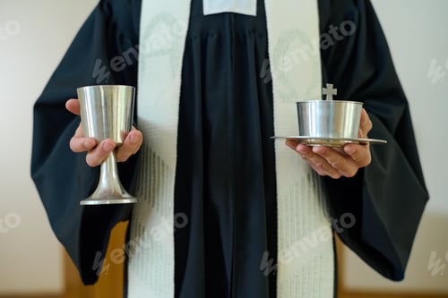 Preview: Hands of Catholic priest in cassock holding cups with wine and unleavened bread
